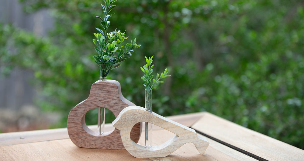 2 wooden vases one a bird shape and one an organic pear shape sitting on wooden table outdoors in front of green bushs