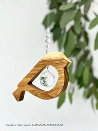 A wooden bird shaped suncatcher made from Tasmanian Blackwood hanging from a wire with a large faceted crystal in the centre and smaller crystal beads on top. Hanging in front of an eucalyptus branch