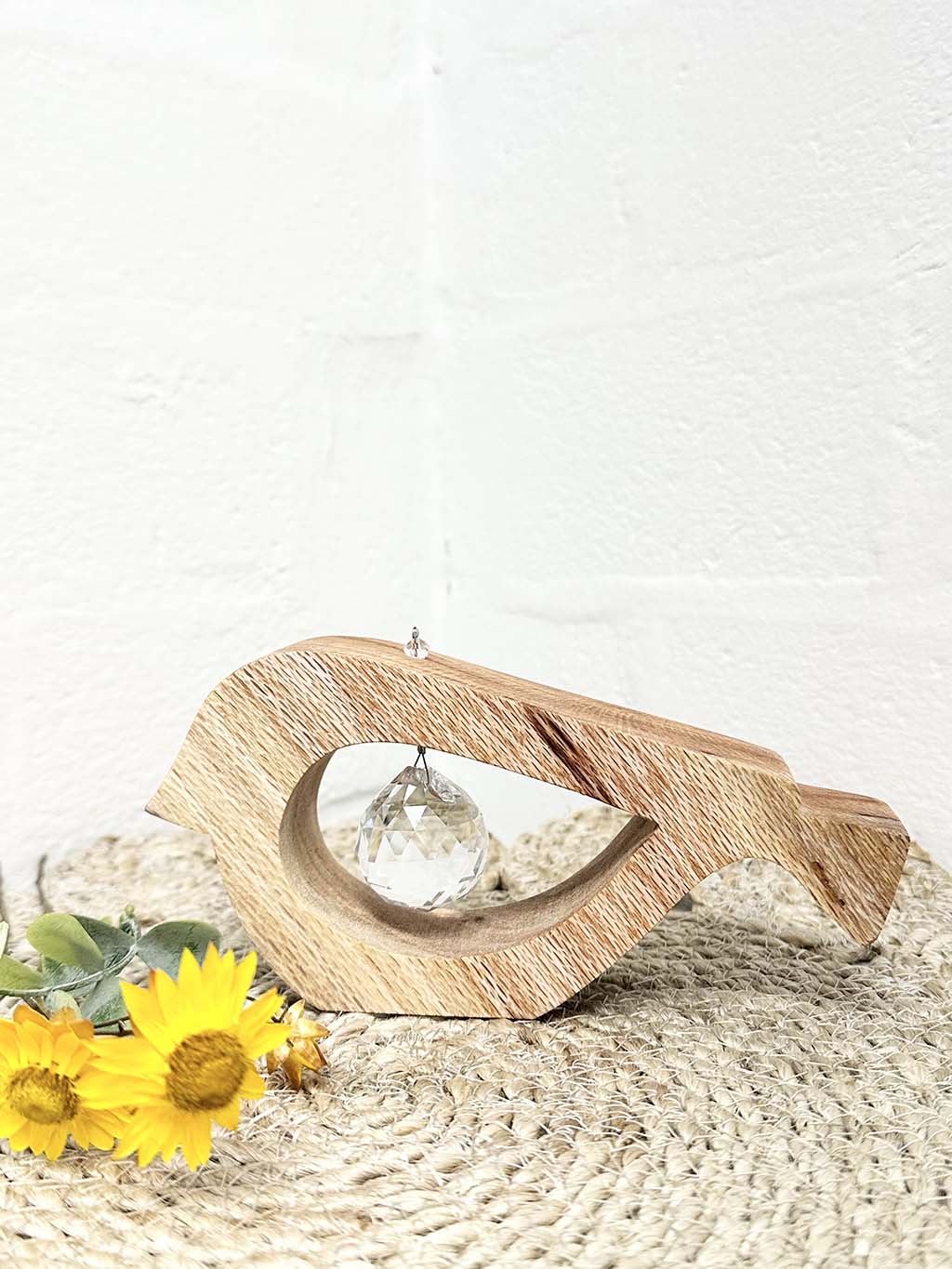 A handmade wooden bird shaped table decoration. The modern shaped bird is cut out from silky oak timber and has a large crystal hanging in the cut out centre shape. There is a yellow paper daisy alongside the bird.