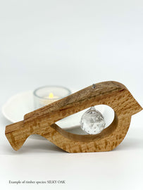 A handmade wooden bird shaped table decoration. The modern shaped bird is cut out from silky oak timber and has a large crystal hanging in the cut out centre shape. There is a tealight candle lit and glowing in the background.