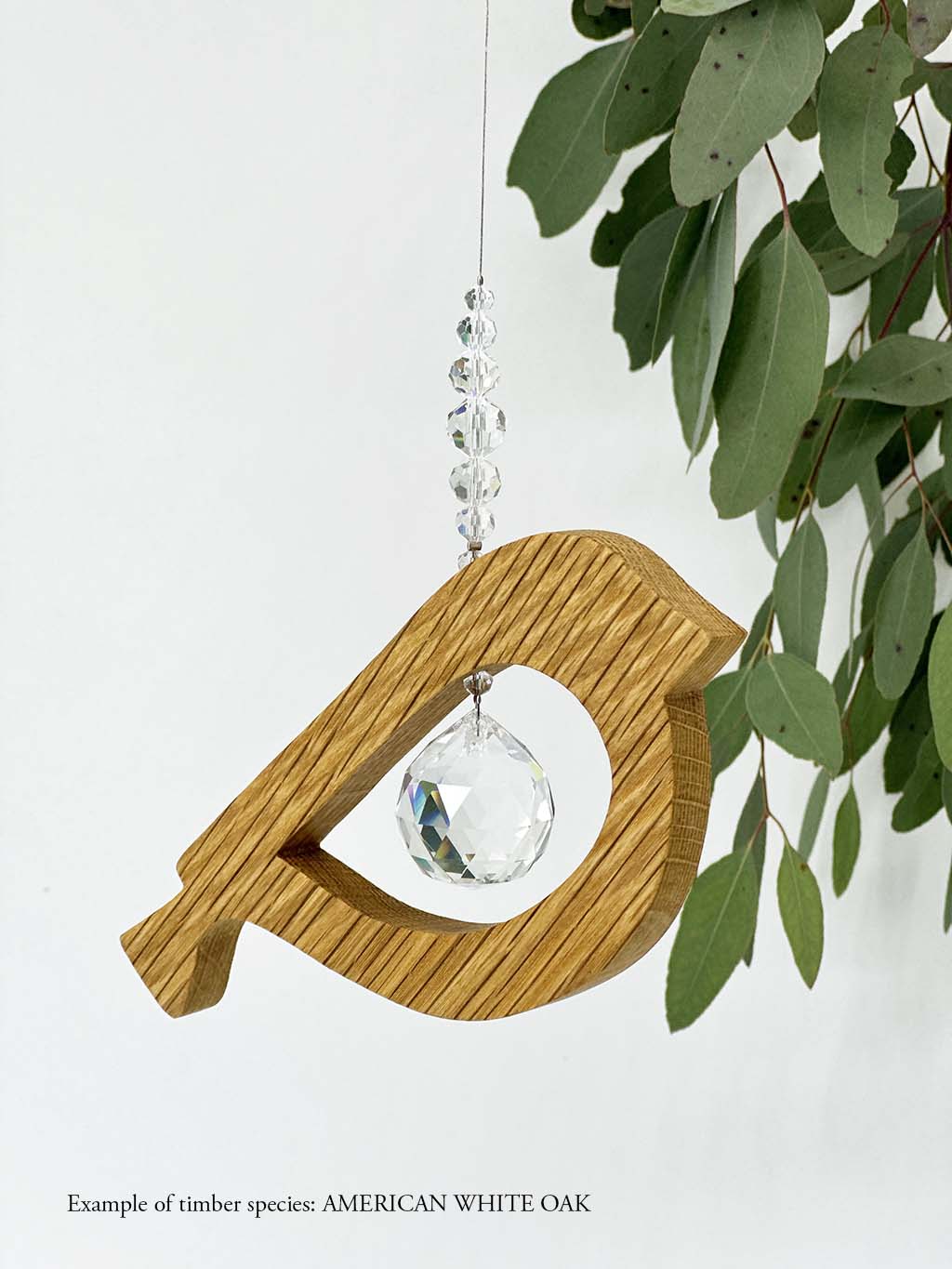 A wooden bird shaped suncatcher made from American White Oak hanging from a wire with a large faceted crystal in the centre and smaller crystal beads on top. Hanging in front of an eucalyptus branch