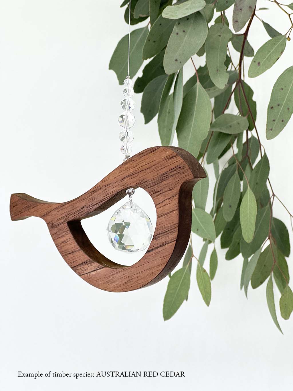 A wooden bird shaped suncatcher made from Australian Red Cedar hanging from a wire with a large faceted crystal in the centre and smaller crystal beads on top. Hanging in front of an eucalyptus branch