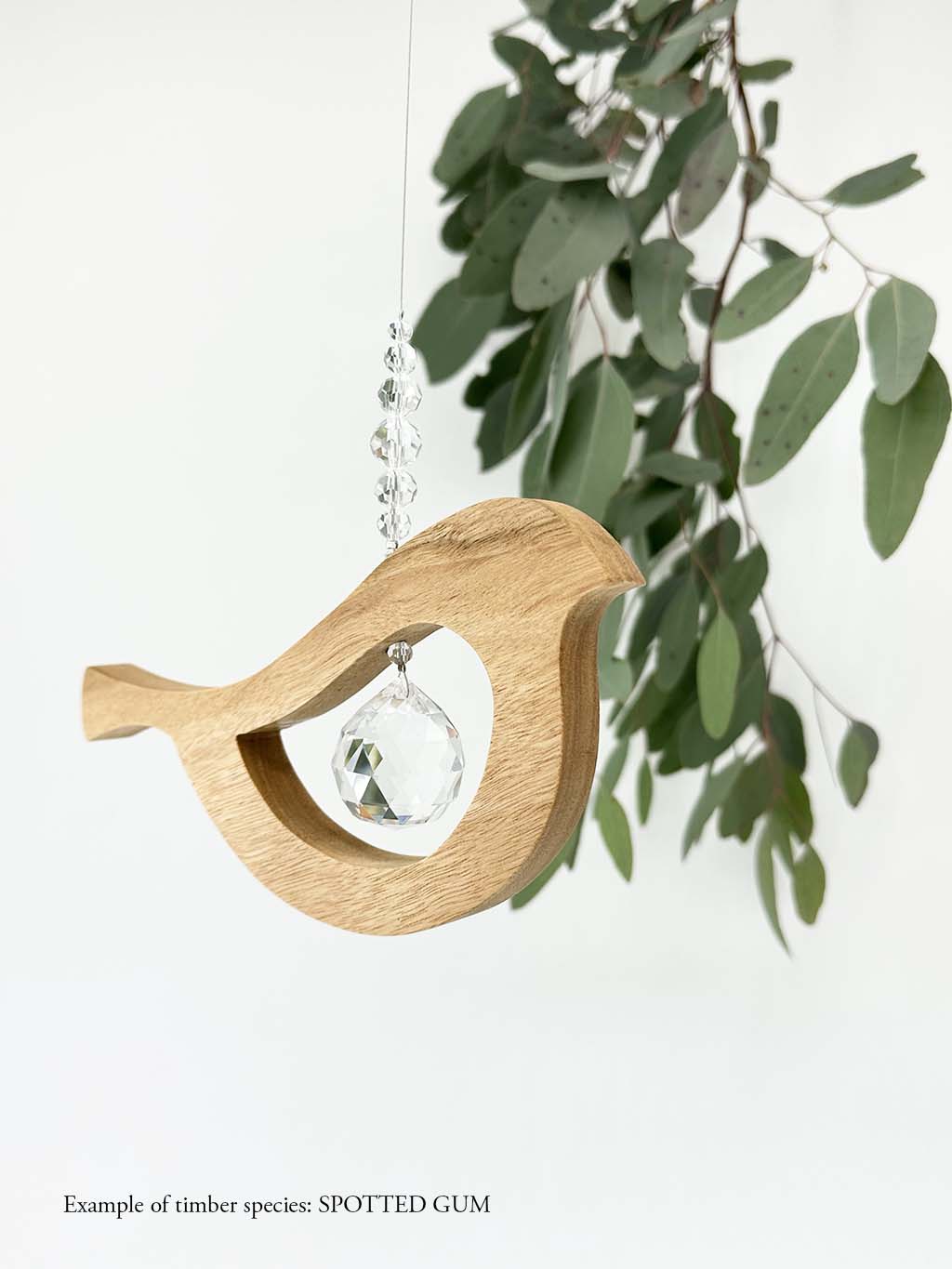 A wooden bird shaped suncatcher made from Spotted Gum hanging from a wire with a large faceted crystal in the centre and smaller crystal beads on top. Hanging in front of an eucalyptus branch