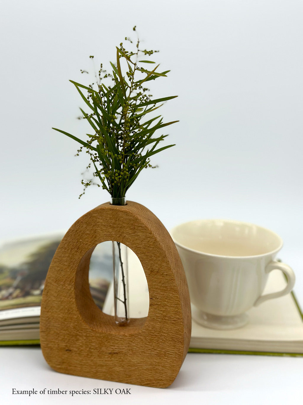 A wooden dome shaped vase with a glass test tube containing a jonquil and wattle sprig made from Silky Oak timber with book and teacup