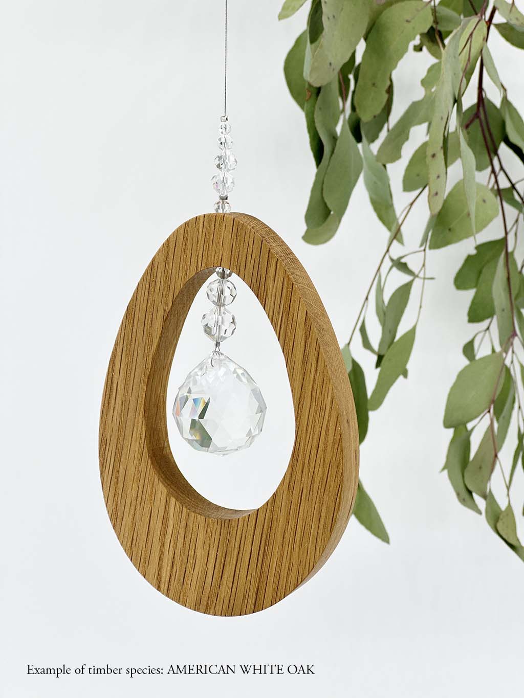 A wooden oval shaped suncatcher made from American White Oak hanging from a wire with a large faceted crystal in the centre and smaller crystal beads on top. Hanging in front of an eucalyptus branch