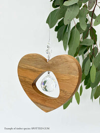A wooden heart shaped suncatcher made from Spotted Gum hanging from a wire with a large faceted crystal in the centre and smaller crystal beads on top. Hanging in front of an eucalyptus branch