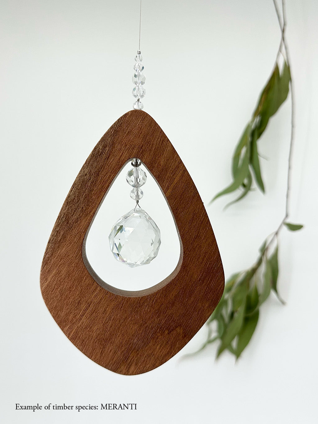 A wooden kite shaped suncatcher made from Meranti hanging from a wire with a large faceted crystal in the centre and smaller crystal beads on top. Hanging in front of an eucalyptus branch