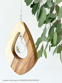 A wooden kite shaped suncatcher made from Tasmanian Blackwood hanging from a wire with a large faceted crystal in the centre and smaller crystal beads on top. Hanging in front of an eucalyptus branch