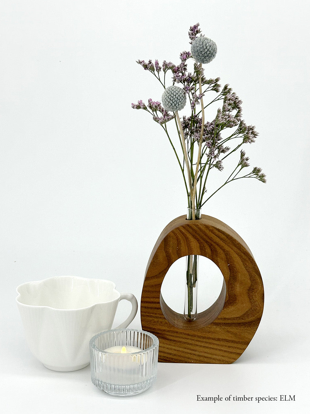 A wooden oval shaped vase with a glass test tube containing a flower and billy button sprig made from Elm timber near a teacup and candle