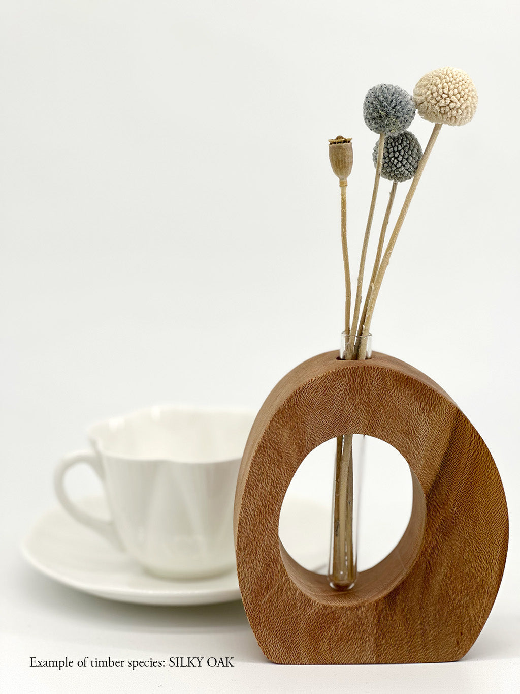 A wooden oval shaped vase with a glass test tube containing dried flower sprig made from Silky Oak timber near a teacup
