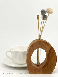 A wooden oval shaped vase with a glass test tube containing dried flower sprig made from Silky Oak timber near a teacup