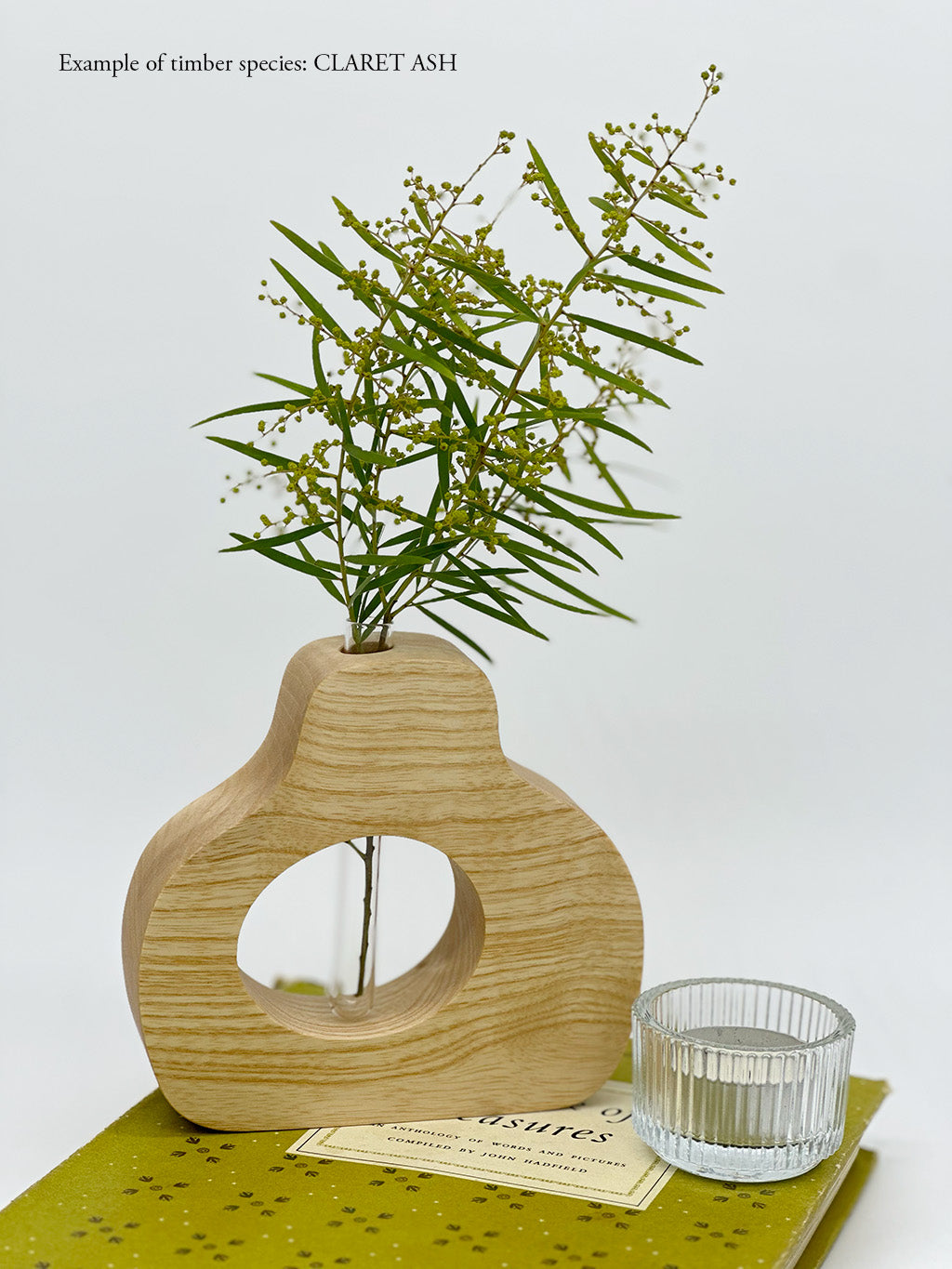 A wooden pear shaped vase with a glass test tube containing Wattle sprig made from Claret Ash timber on a book and beside a candle