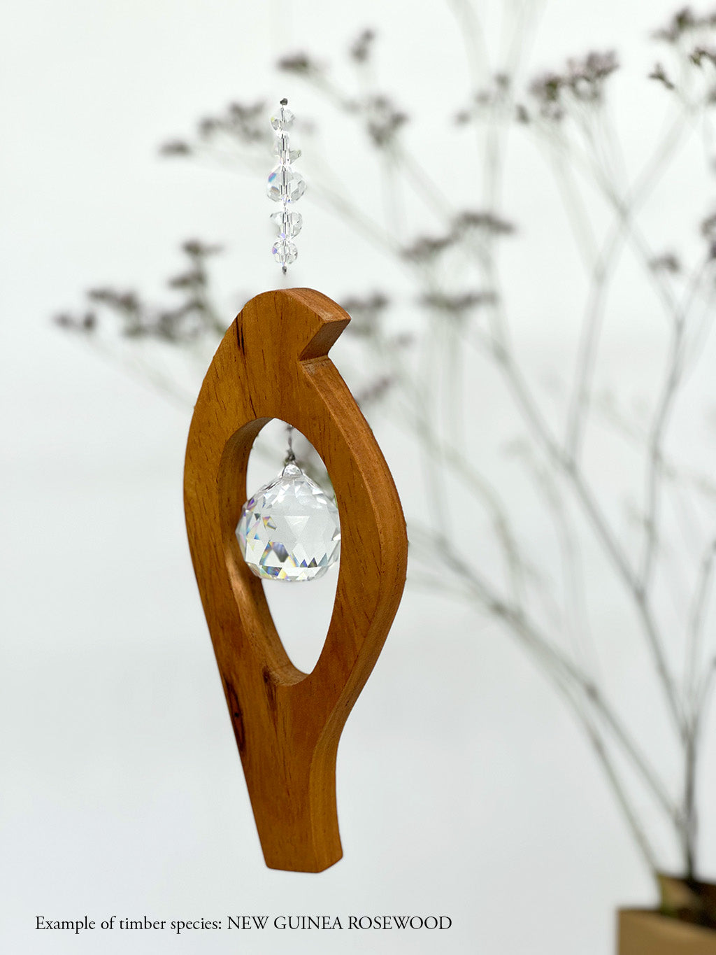 A wooden bird shaped suncatcher made from New Guinea Rosewood hanging from a wire with a large faceted crystal in the centre and smaller crystal beads on top.