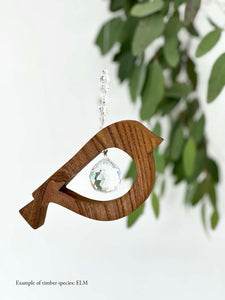 A wooden bird shaped suncatcher made from Elm hanging from a wire with a large faceted crystal in the centre and smaller crystal beads on top. Hanging in front of an eucalyptus branch
