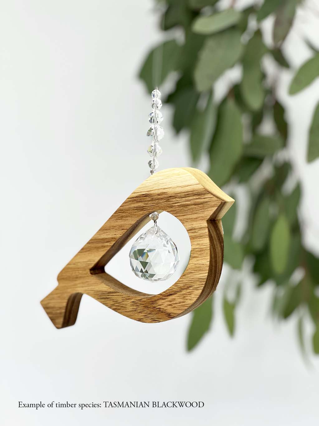 A wooden bird shaped suncatcher made from Tasmanian Blackwood hanging from a wire with a large faceted crystal in the centre and smaller crystal beads on top. Hanging in front of an eucalyptus branch