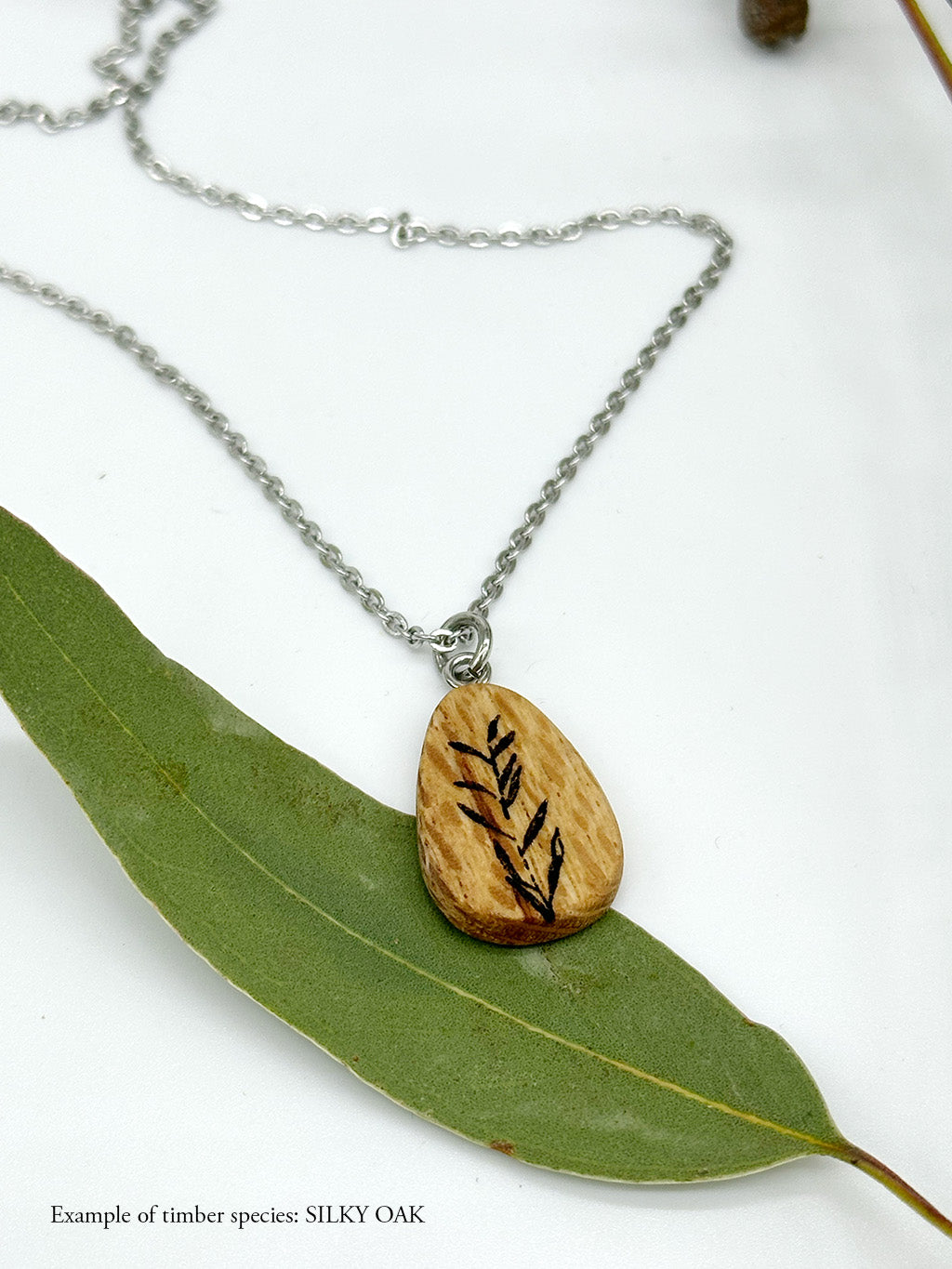 Wooden Silky Oak pendant almond shape with botanical illustration lying on a eucalyptus leaf