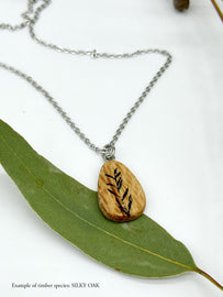 Wooden Silky Oak pendant almond shape with botanical illustration lying on a eucalyptus leaf