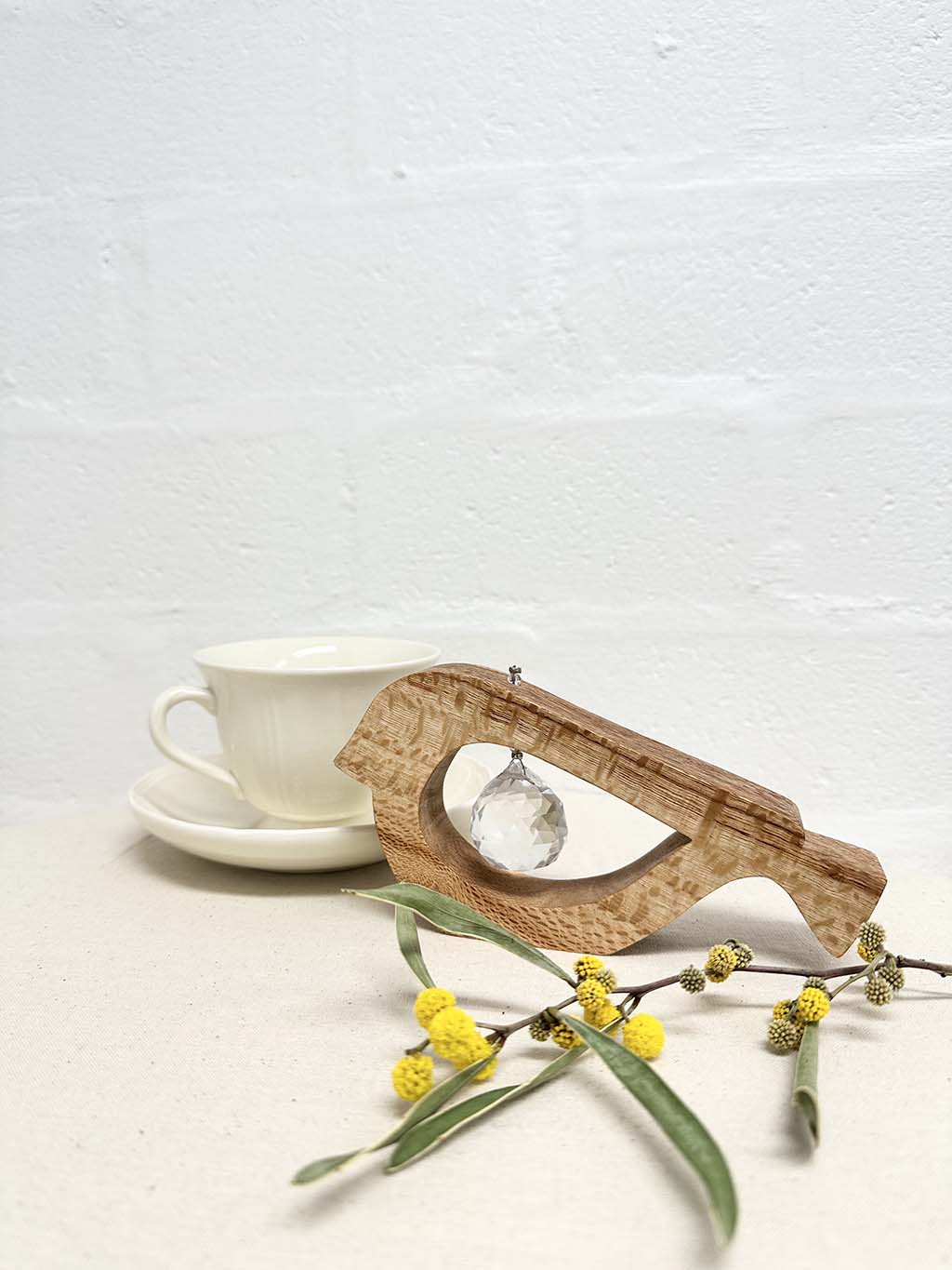 A handmade wooden bird shaped table decoration. The modern shaped bird is cut out from silky oak timber and has a large crystal hanging in the cut out centre shape. There is a teacup and saucer in the background and a sprig of wattle in the foreground.