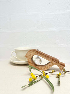 A handmade wooden bird shaped table decoration. The modern shaped bird is cut out from silky oak timber and has a large crystal hanging in the cut out centre shape. There is a teacup and saucer in the background and a sprig of wattle in the foreground.