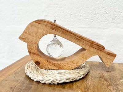 A handmade wooden bird shaped table decoration. The modern shaped bird is cut out from silky oak timber and has a large crystal hanging in the cut out centre shape. The bird is sitting on a raffia placemat.