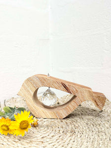 A handmade wooden bird shaped table decoration. The modern shaped bird is cut out from silky oak timber and has a large crystal hanging in the cut out centre shape. There is a yellow paper daisy alongside the bird.