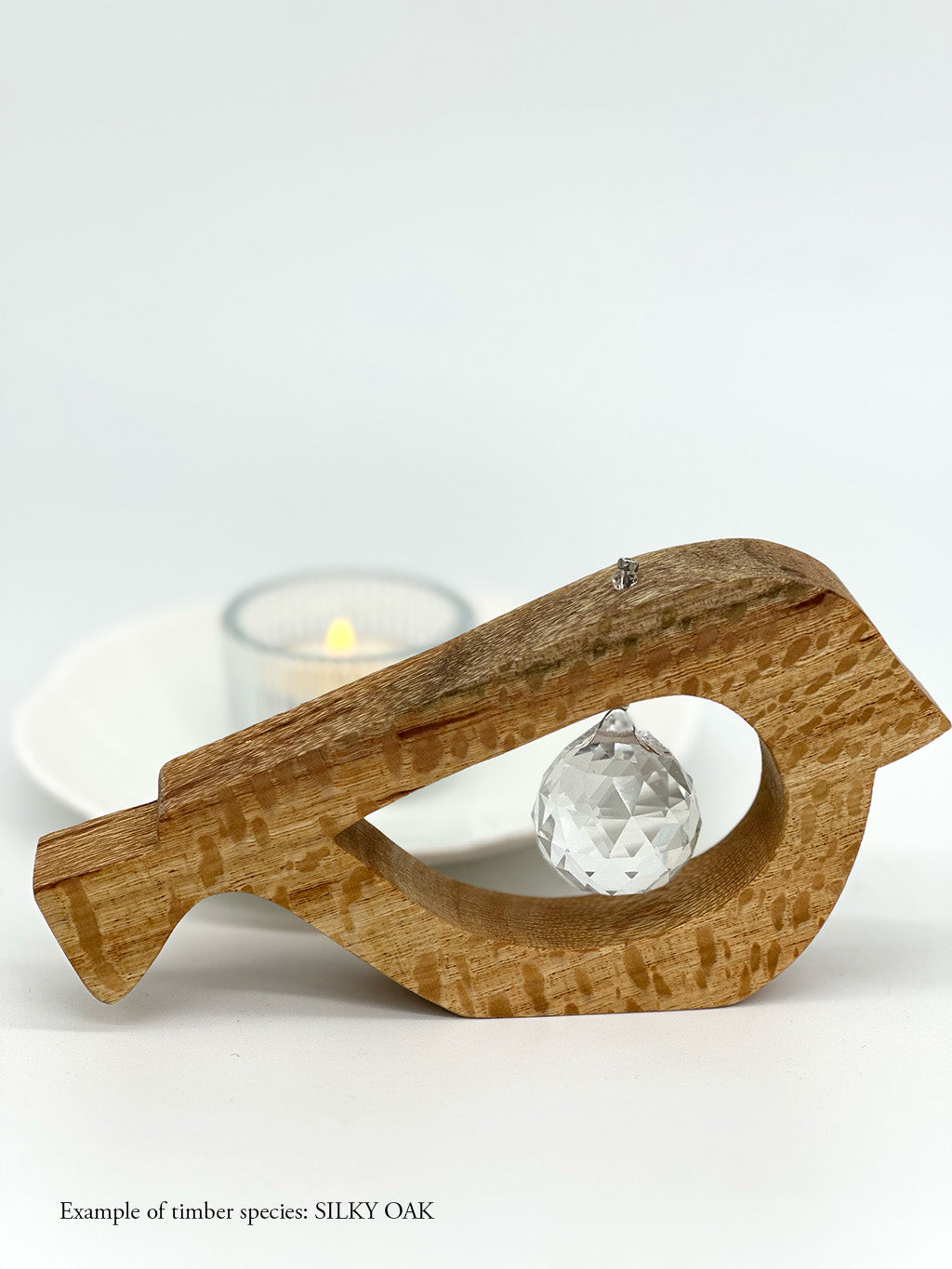 A handmade wooden bird shaped table decoration. The modern shaped bird is cut out from silky oak timber and has a large crystal hanging in the cut out centre shape. There is a tealight candle lit and glowing in the background.