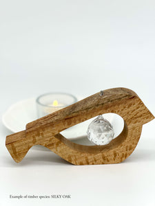 A handmade wooden bird shaped table decoration. The modern shaped bird is cut out from silky oak timber and has a large crystal hanging in the cut out centre shape. There is a tealight candle lit and glowing in the background.