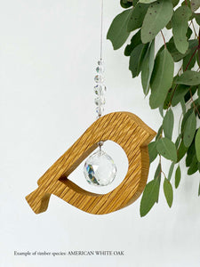 A wooden bird shaped suncatcher made from American White Oak hanging from a wire with a large faceted crystal in the centre and smaller crystal beads on top. Hanging in front of an eucalyptus branch