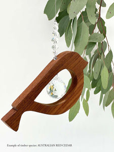 A wooden bird shaped suncatcher made from Australian Red Cedar hanging from a wire with a large faceted crystal in the centre and smaller crystal beads on top. Hanging in front of an eucalyptus branch