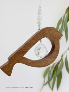 A wooden bird shaped suncatcher made from Meranti hanging from a wire with a large faceted crystal in the centre and smaller crystal beads on top. Hanging in front of an eucalyptus branch