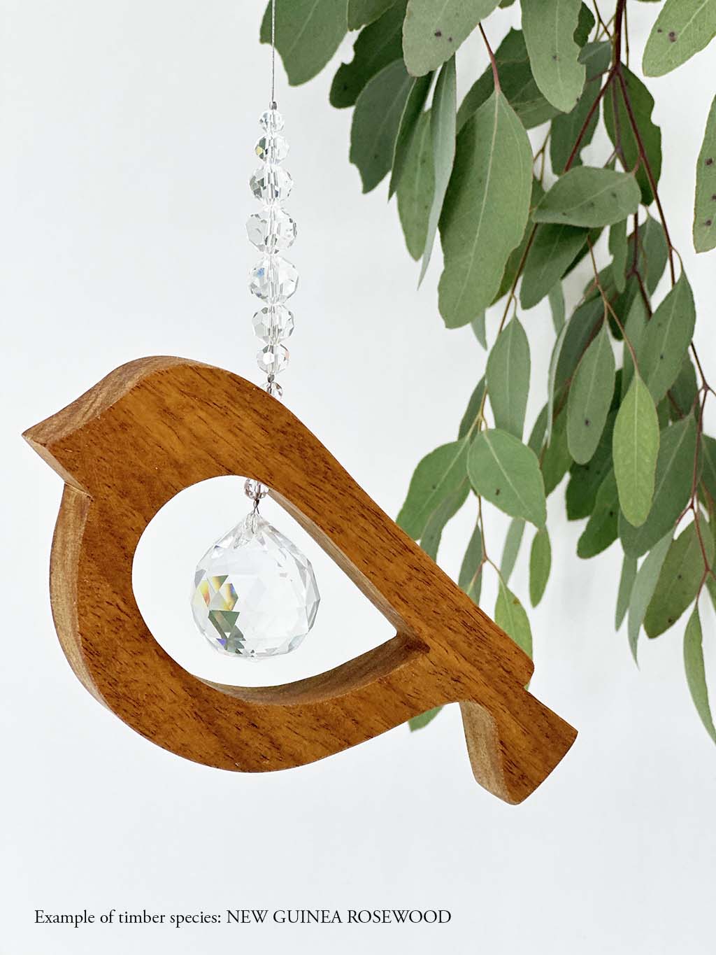 A wooden bird shaped suncatcher made from New Guinea Rosewood hanging from a wire with a large faceted crystal in the centre and smaller crystal beads on top. Hanging in front of an eucalyptus branch