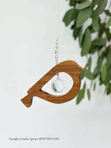 A wooden bird shaped suncatcher made from Spotted Gum hanging from a wire with a large faceted crystal in the centre and smaller crystal beads on top. Hanging in front of an eucalyptus branch