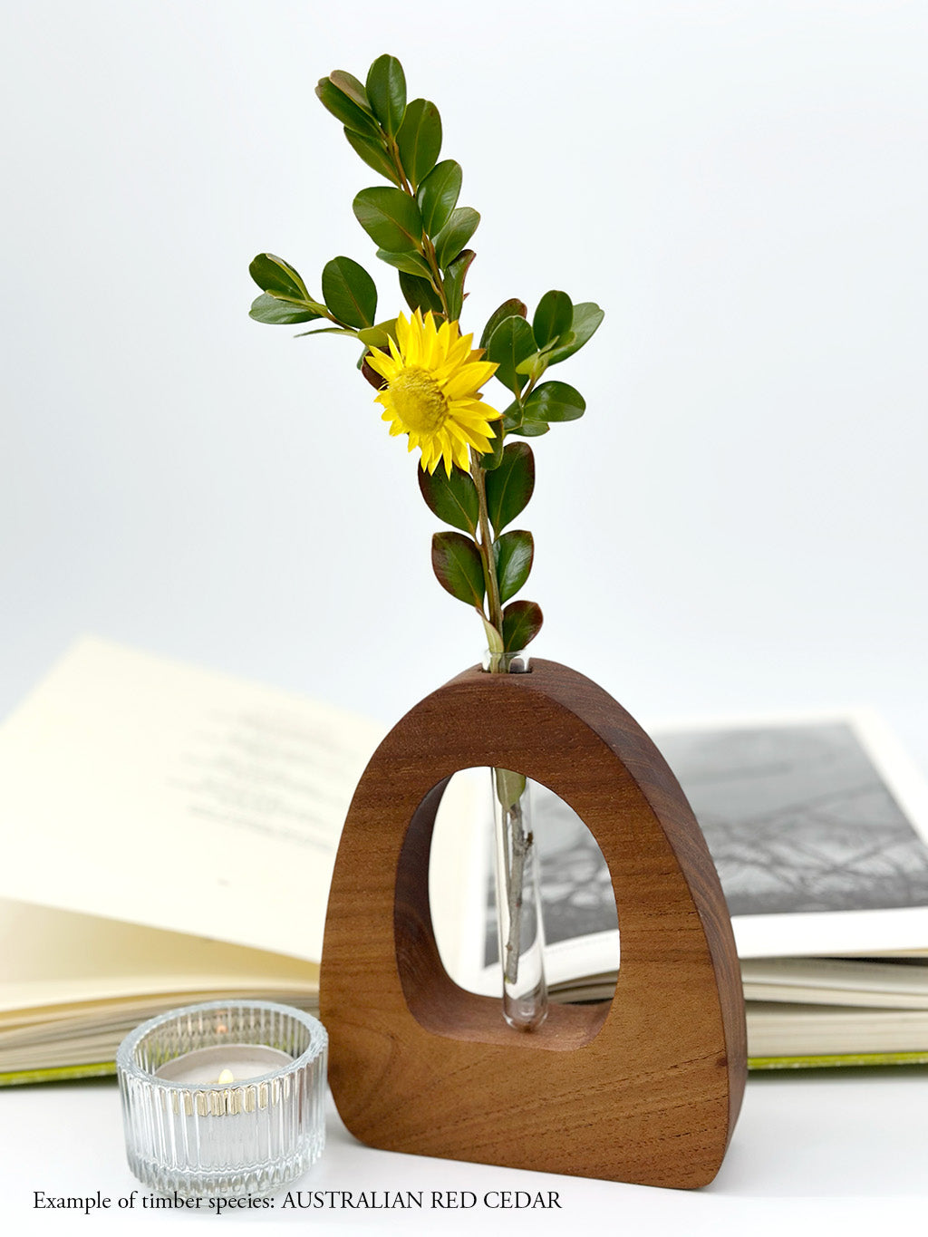 A wooden dome shaped vase with a glass test tube containing a yellow daisy sprig made from Australian Red Cedar timber with an open book and candle