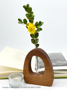 A wooden dome shaped vase with a glass test tube containing a yellow daisy sprig made from Australian Red Cedar timber with an open book and candle