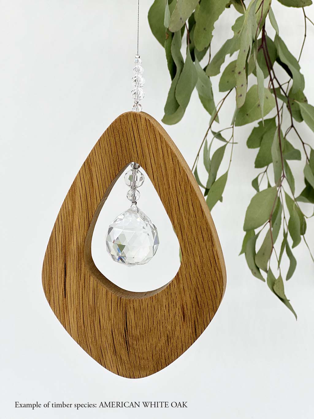 A wooden kite shaped suncatcher made from American White Oak hanging from a wire with a large faceted crystal in the centre and smaller crystal beads on top. Hanging in front of an eucalyptus branch