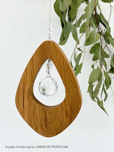 A wooden kite shaped suncatcher made from American White Oak hanging from a wire with a large faceted crystal in the centre and smaller crystal beads on top. Hanging in front of an eucalyptus branch