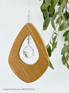 A wooden kite shaped suncatcher made from Spotted Gum hanging from a wire with a large faceted crystal in the centre and smaller crystal beads on top. Hanging in front of an eucalyptus branch