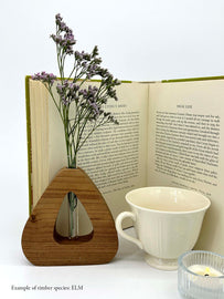 A wooden triangle shaped vase with a glass test tube containing a plant sprig made from Elm timber near a book teacup and candle