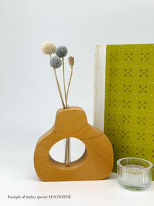 A wooden pear shaped vase with a glass test tube containing Billy Button sprig made from Huon Pine timber near a book and candle